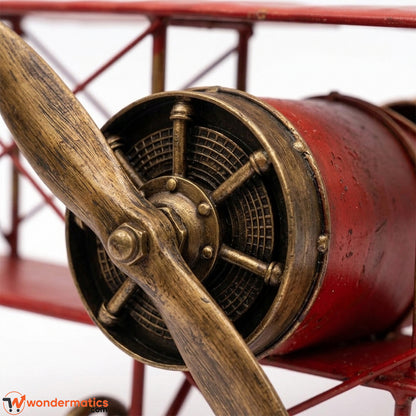 Close-up of a vintage-style biplane model with a red body, brass-colored engine details, and a wooden propeller blade against a white background.