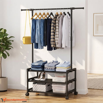 A black metal rolling garment rack with a top bar holding jeans, shirts, and a plaid button-up, plus two lower storage shelves with folded clothes and storage bins, shown in a bright room with a plant and wall art.