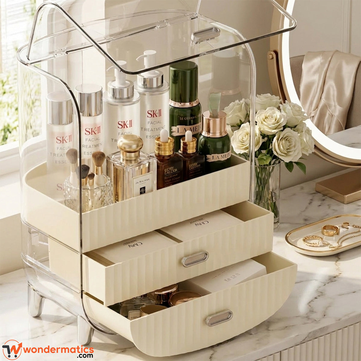 A clear acrylic and cream-colored rolling makeup organizer filled with skincare and cosmetic products, set on a marble vanity next to white roses and a ring light.