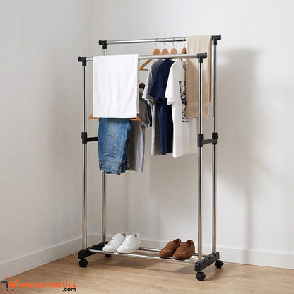 A silver metal clothes rack on wheels with black plastic joints and feet, shown in a room corner with clothes hanging on hangers, a white towel, and shoes on the bottom shelf.