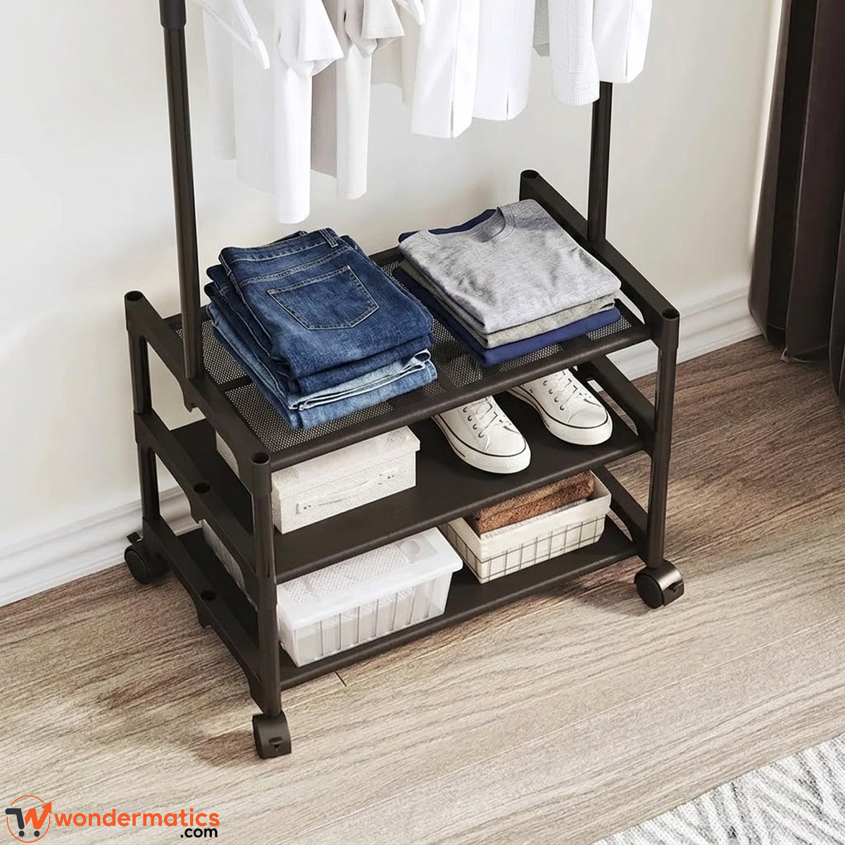 A black rolling three-tier clothing organizer with a top rack holding white shirts, shelves folded in blue jeans, a gray sweatshirt, white sneakers, and storage bins, set on a wood floor against a white wall.