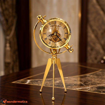 A gold-finished mechanical globe clock on a tripod stand, displayed on a polished dark wood table with a marble inlay. The clock features a transparent glass dome showing the inner gears and Roman numerals on the face.