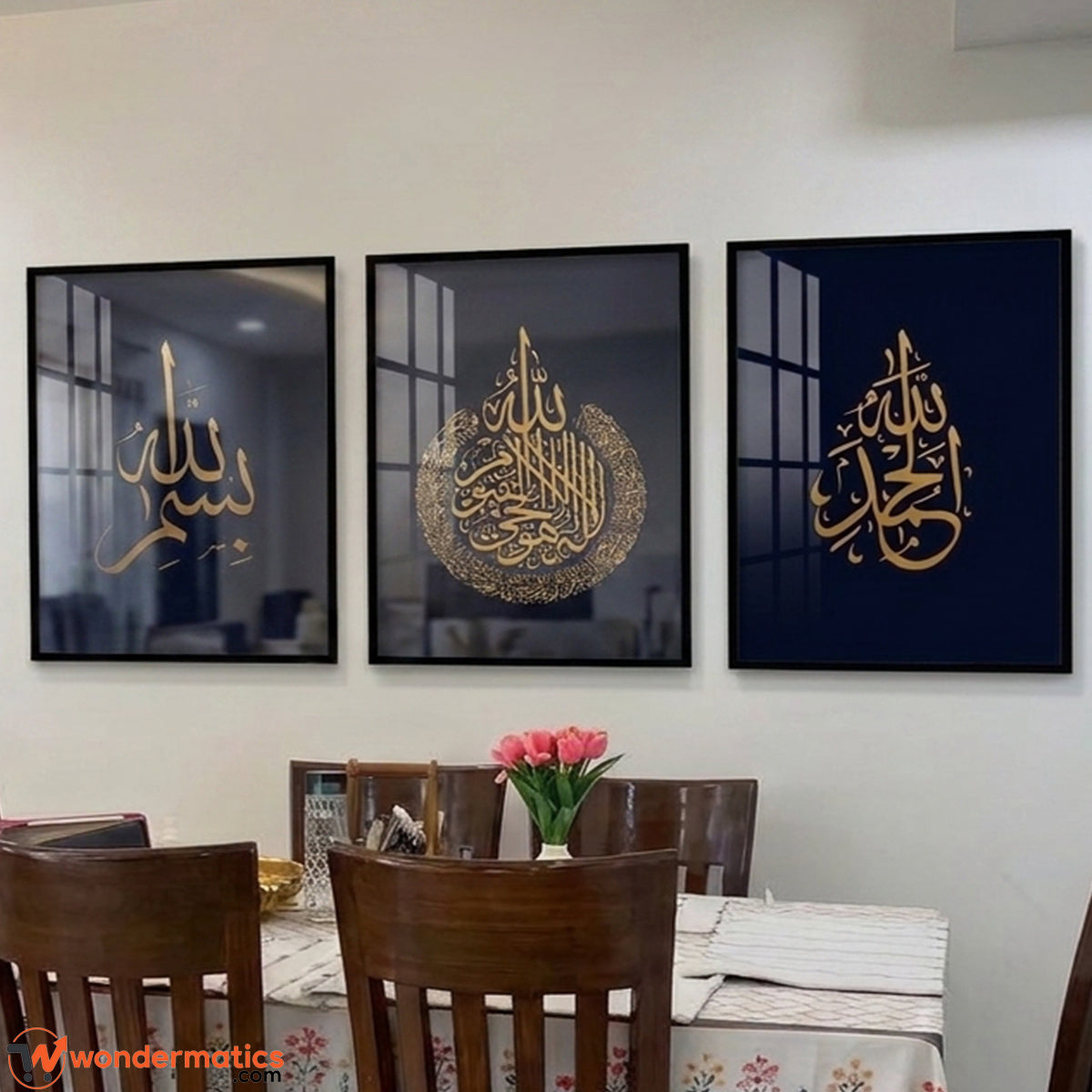 Three framed Islamic wall art prints with gold Arabic calligraphy hang on a light beige wall above a dining table with wooden chairs and a pink flower vase.