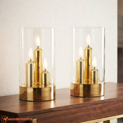 Two gold and clear glass candle holders with lit led flameless candles, displayed on a dark wooden table in a softly blurred indoor setting.