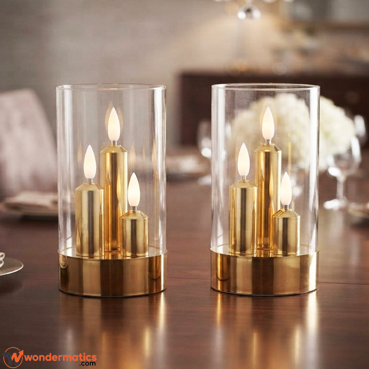 Two gold and clear glass candle holders with lit flameless candles, displayed on a dark wooden table in a softly blurred indoor setting.